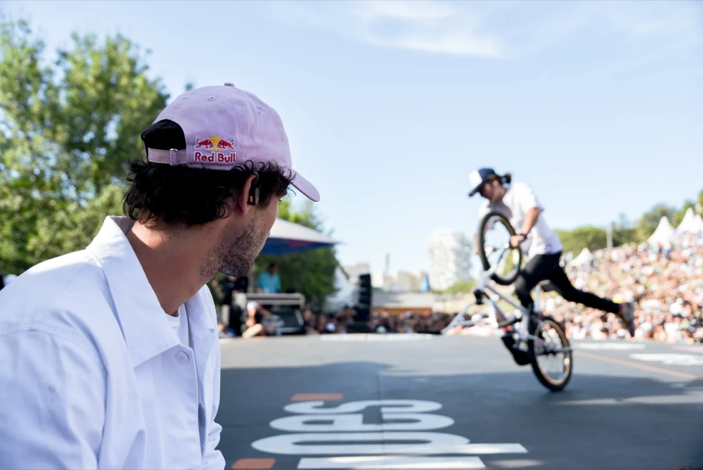 Rider BMX exécutant un trick en flat, observé depuis l’avant-plan par un spectateur avec casquette rose.