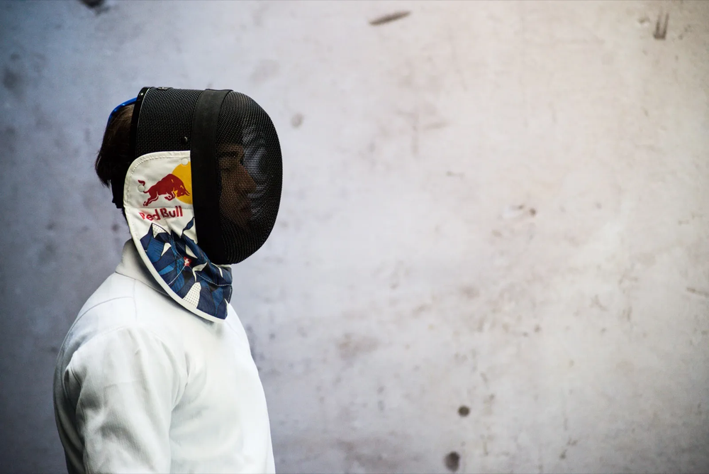 Portrait d’un escrimeur sponsorisé par Red Bull, en tenue blanche et masque d’escrime, sur fond neutre.