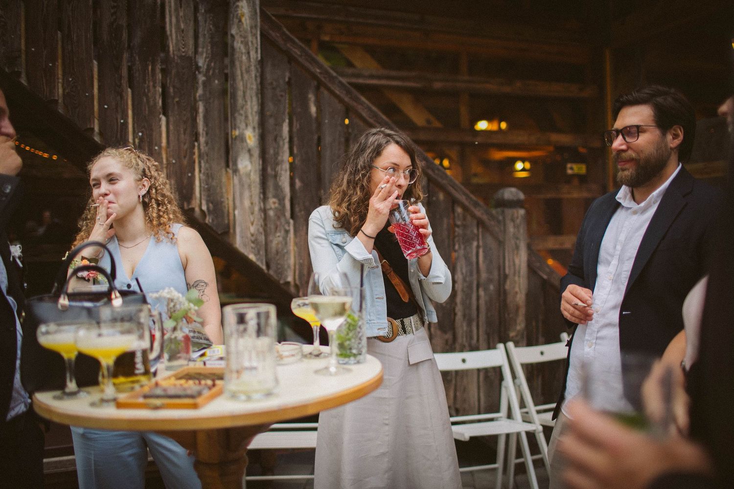 Wedding guests gather on a rustic wooden porch enjoying drinks and conversation during a reception celebration.