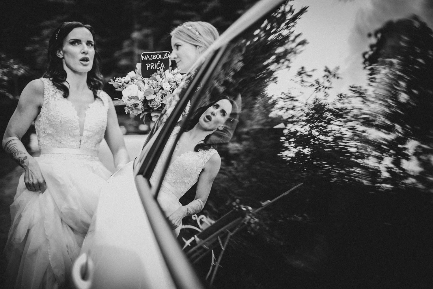 Black and white photo capturing candid moments through a car window at a wedding.