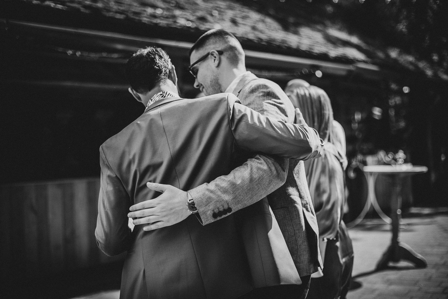 Three people embrace in an emotional black and white moment during an outdoor gathering.