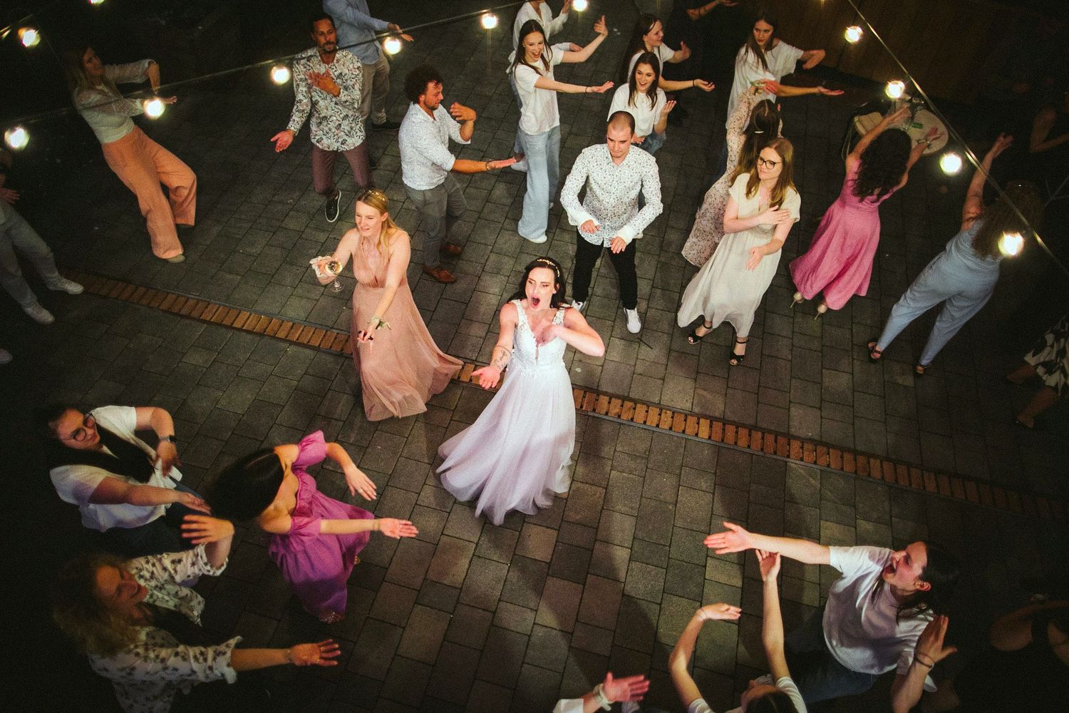 Aerial view of guests dancing in a circle around the newlyweds during an outdoor reception.