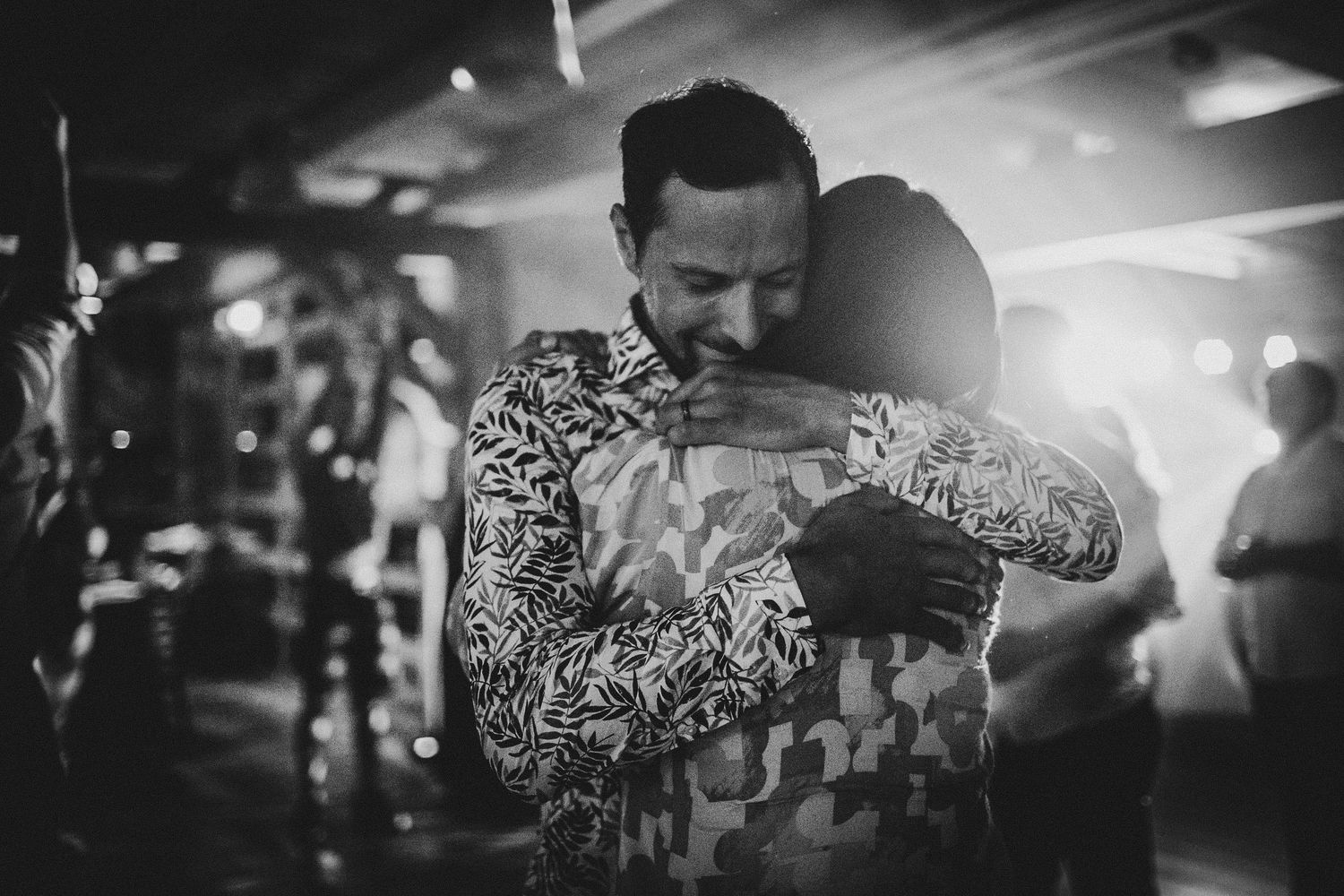 An individual in a patterned shirt holds a mobile device while standing in a dimly lit club or event space.