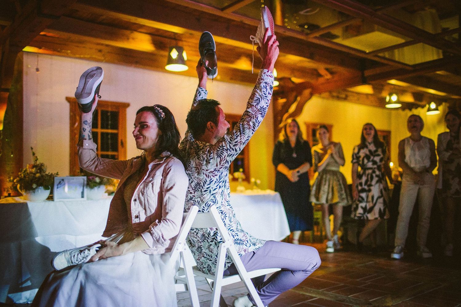 People celebrating during a wedding reception game in a rustic venue with warm lighting and wooden beams.
