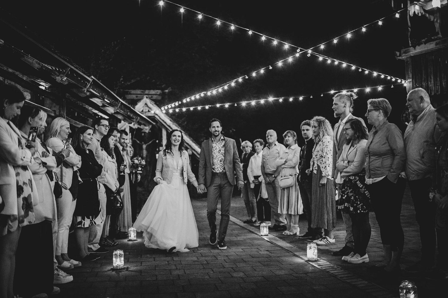A wedding party gathers under twinkling string lights at night as guests line up holding candles along a paved walkway.