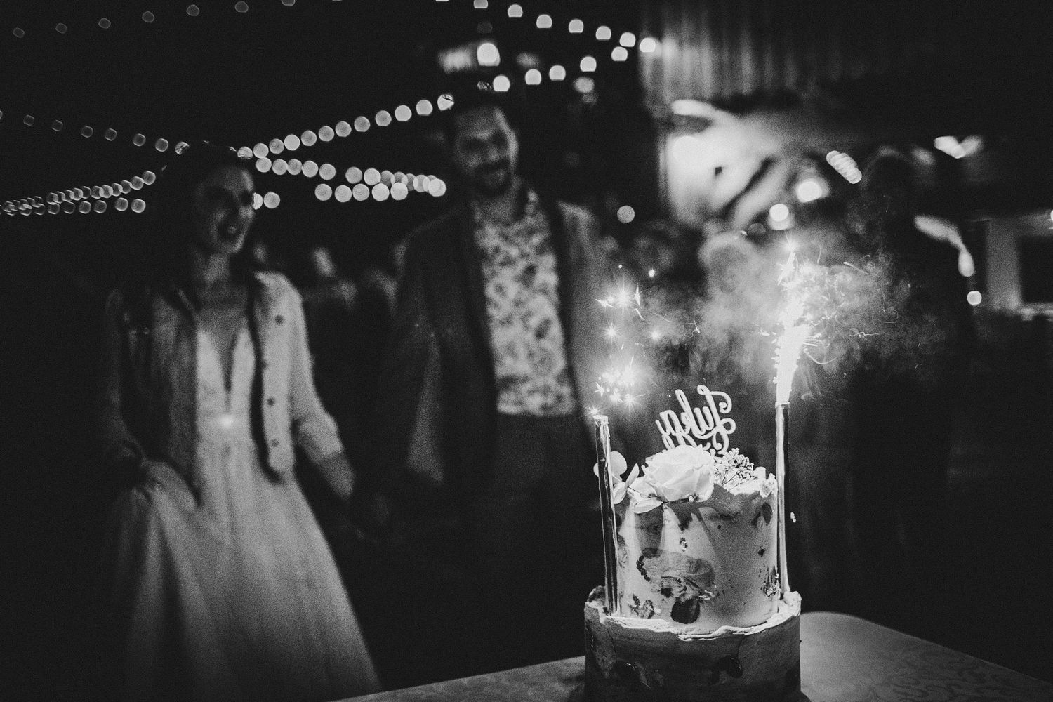 Black and white wedding reception photo showing a glowing sparkler-topped cake with string lights and guests in the background.