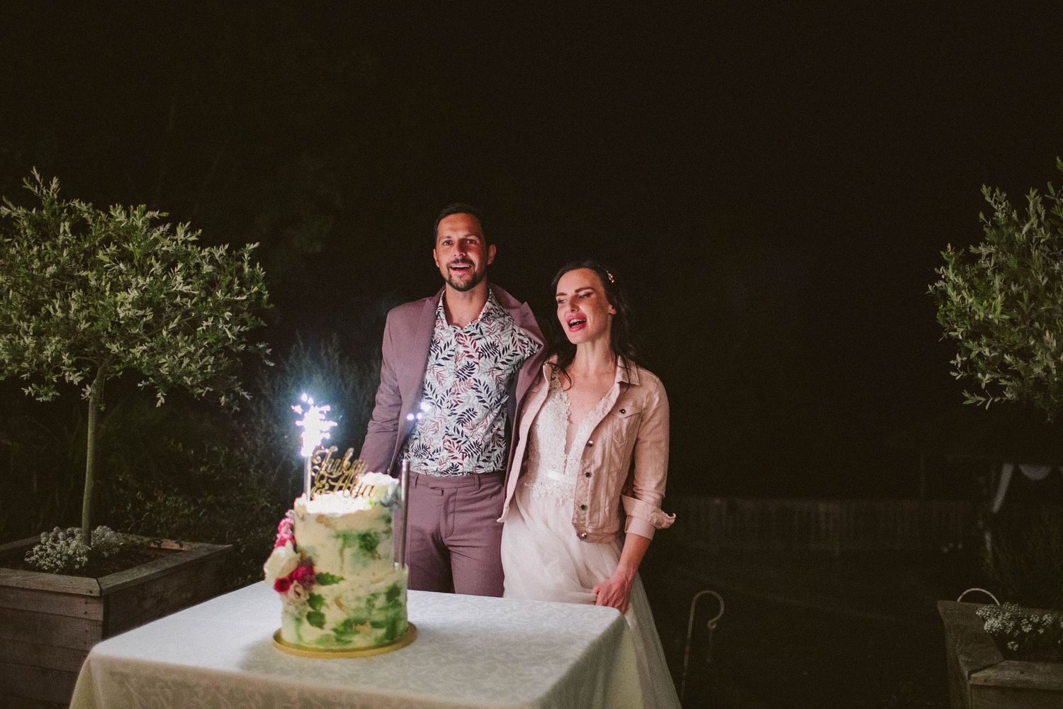 A couple stands beside their decorated green and white wedding cake at an outdoor nighttime reception with sparklers.