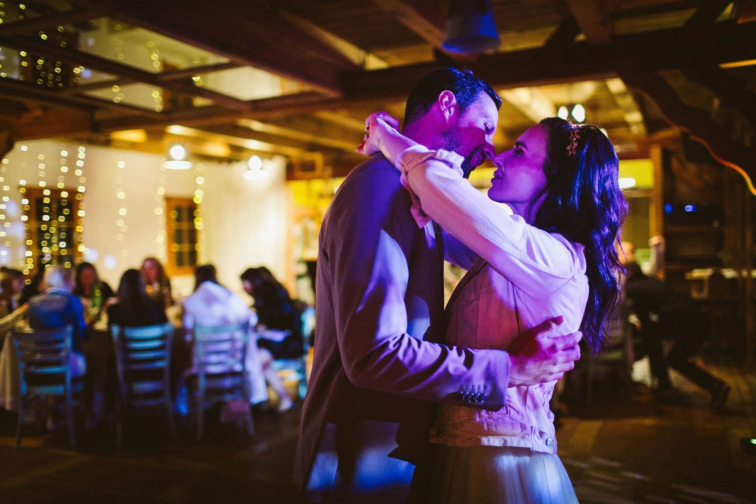 A romantic first dance at a dimly lit wedding reception venue with purple lighting and guests seated in background.