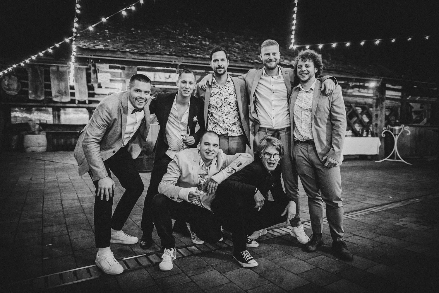 A black and white photo of a group of six people posing together outdoors under string lights at night.