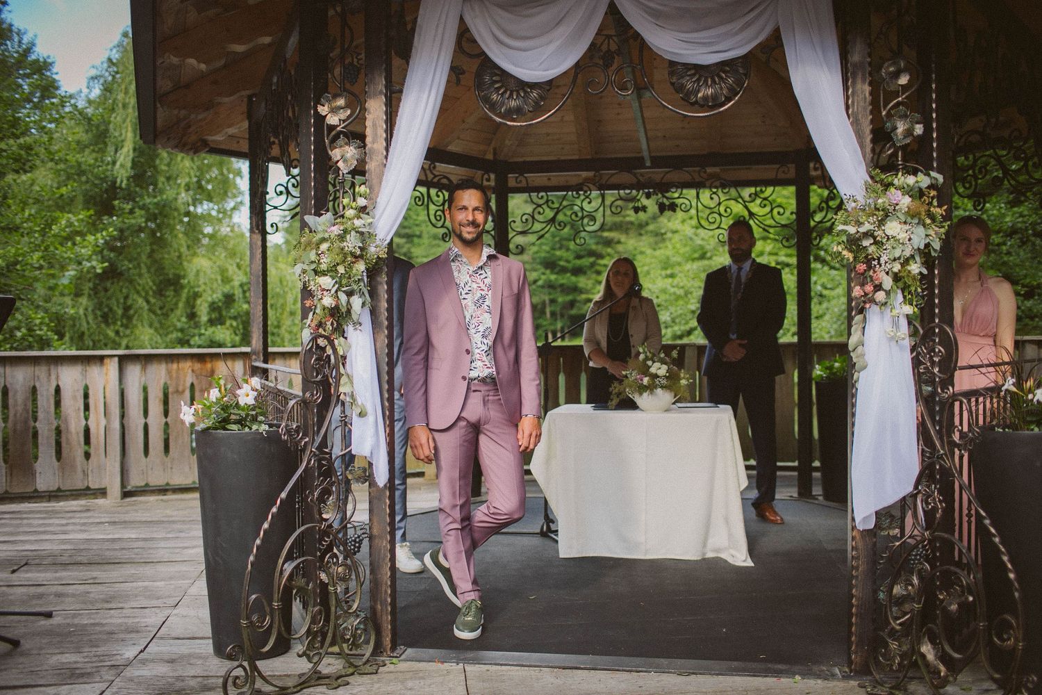 Person in a pink suit stands under a decorated gazebo during an outdoor ceremony.