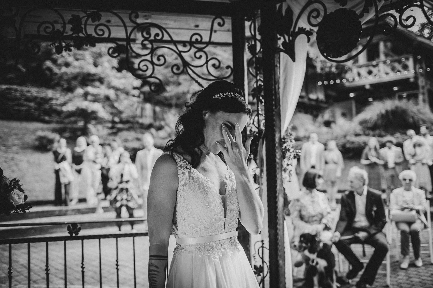 Black and white photo of someone in a wedding dress standing under an ornate gazebo.