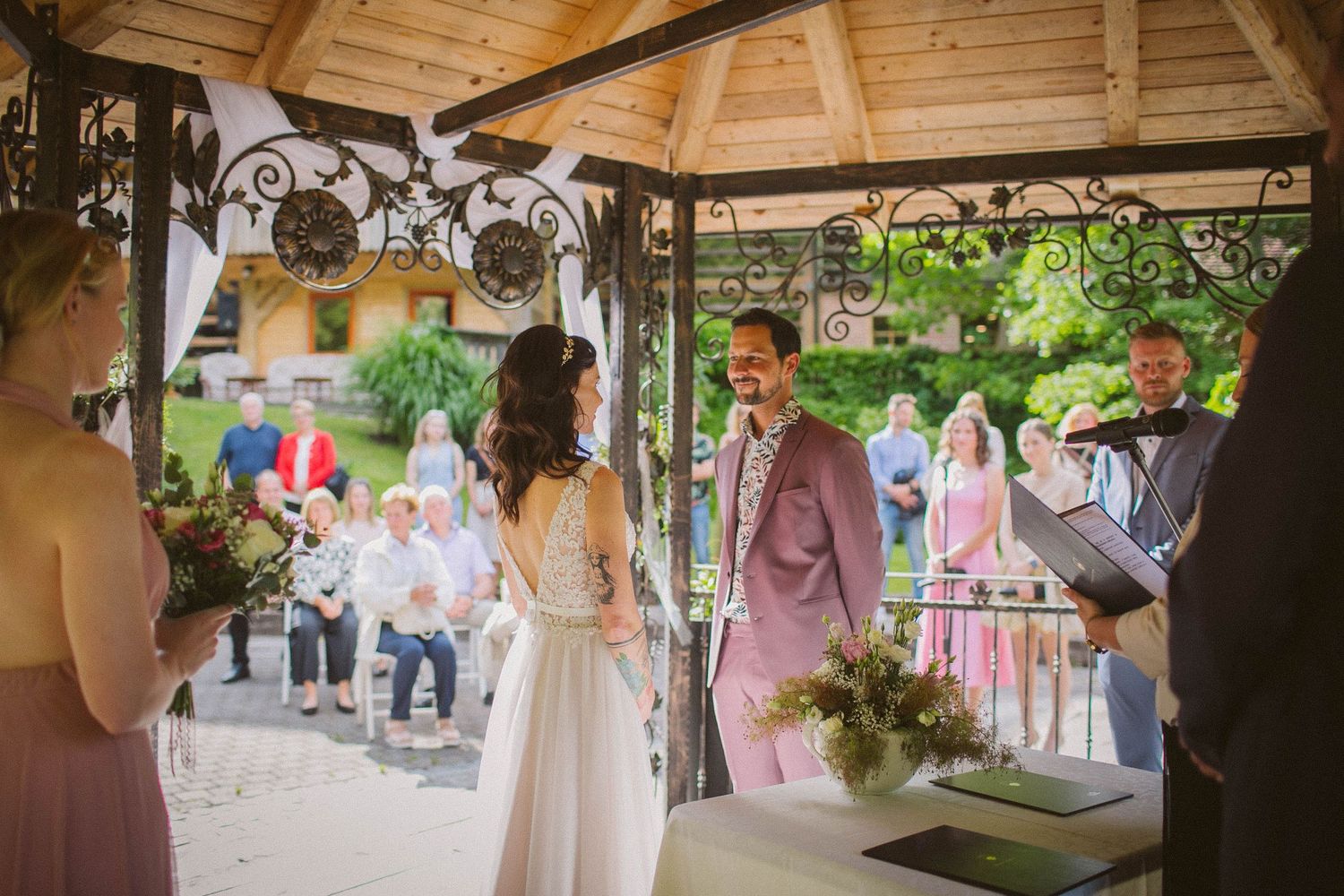 An intimate outdoor wedding ceremony taking place under a wooden gazebo with decorative lights.