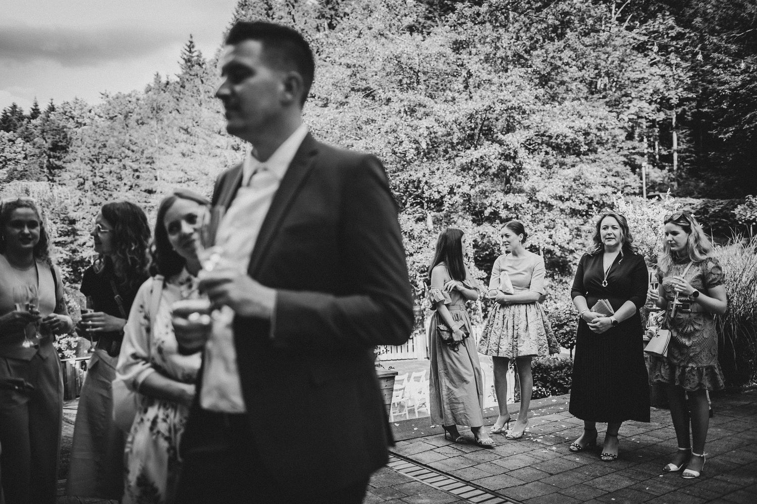 Black and white photo of guests gathered for outdoor wedding celebration in wooded setting.