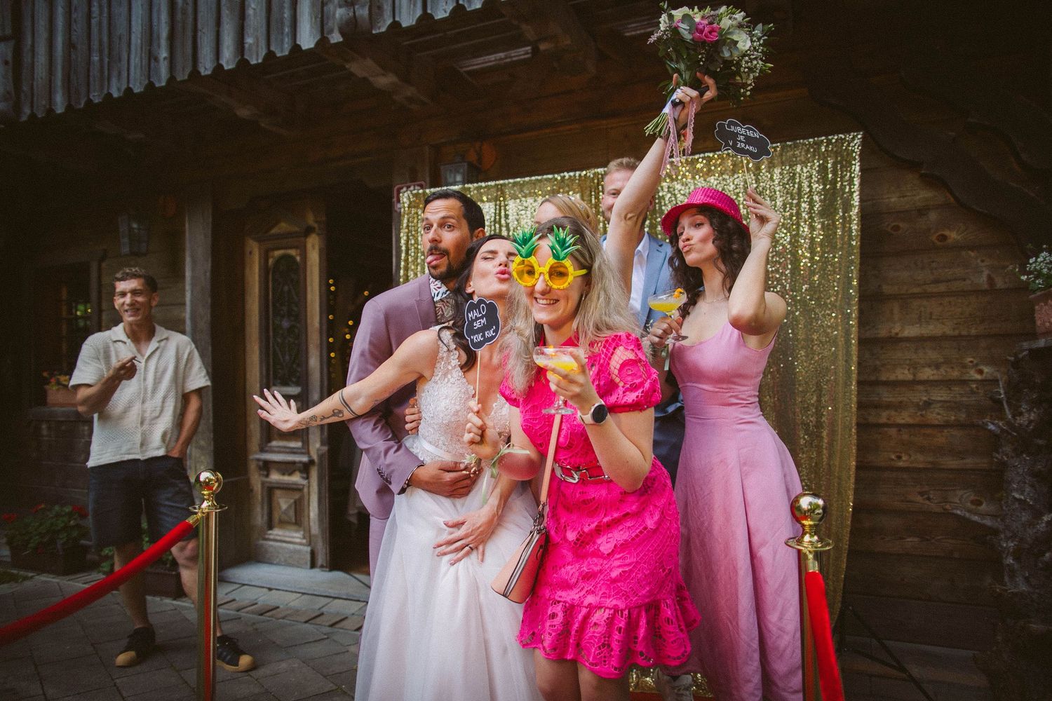 Wedding party poses joyfully with bright pink and pastel dresses against rustic backdrop.