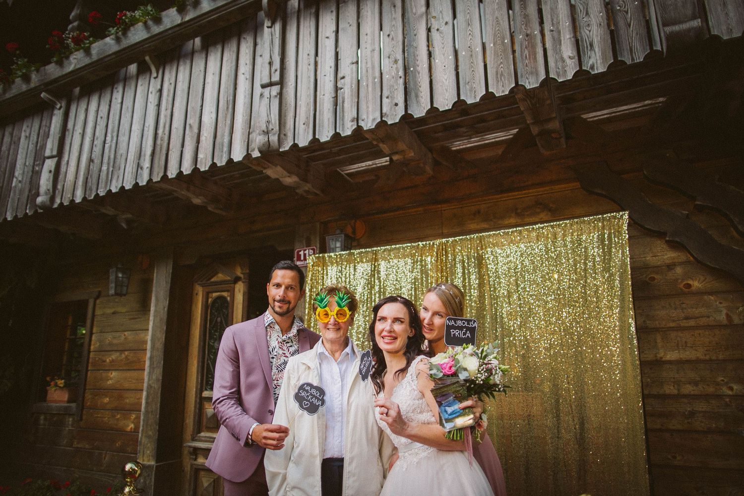 Couple poses with guest in pink suit against gold sequin backdrop at rustic venue.