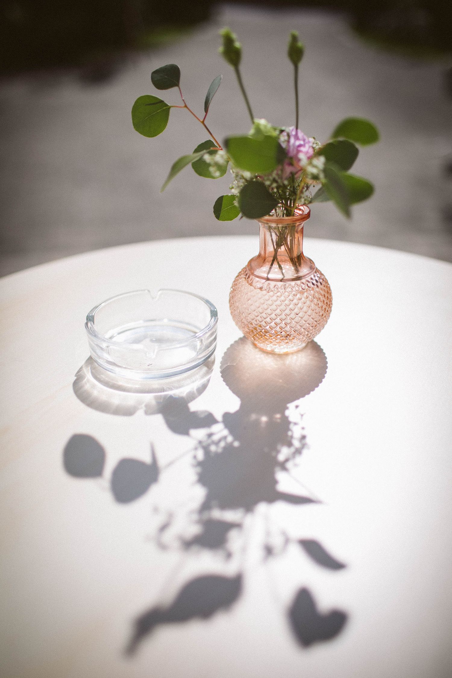 Pink glass vase with delicate flowers casting shadows on white surface in natural light.