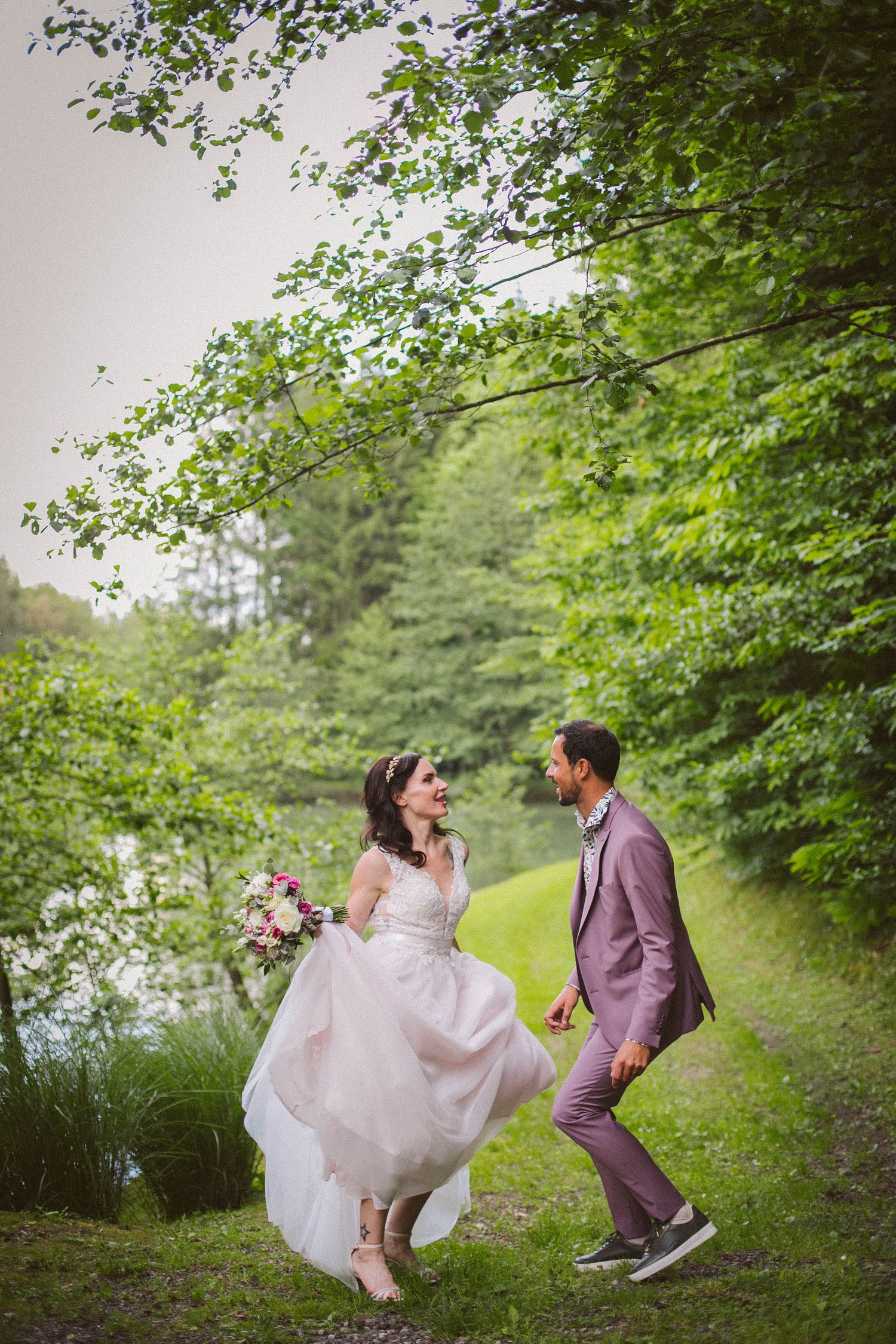 Couple in wedding attire playfully walking on a lush green forest path.