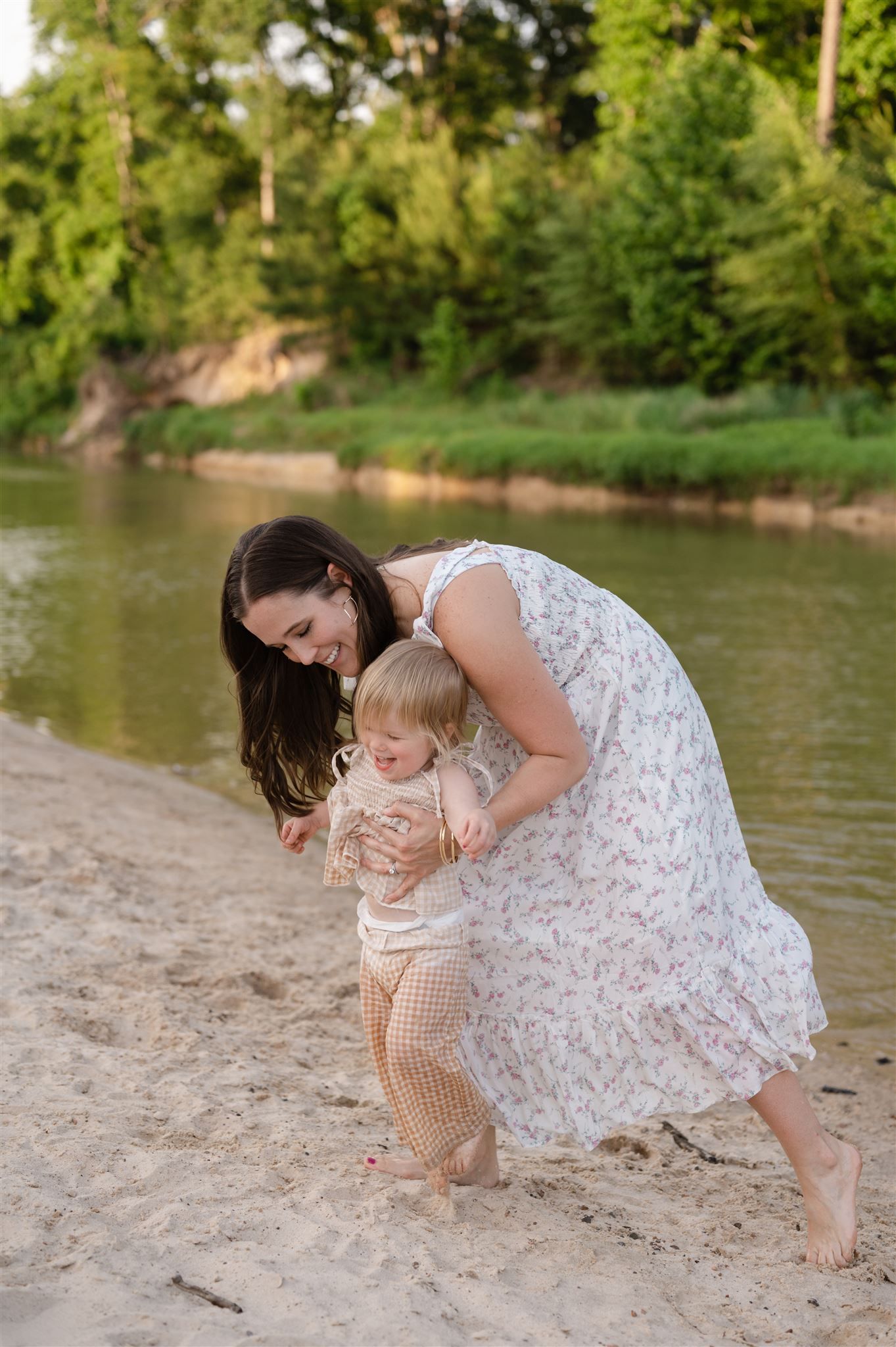 A tender moment shared between two figures in white dresses on a sandy beach by a tranquil river on a summer day.