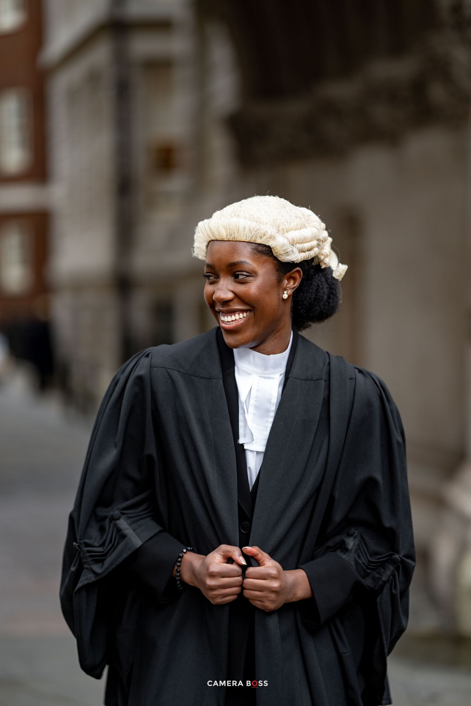 A legal professional in traditional black court robes and white barrister's wig smiles while standing outside a building.