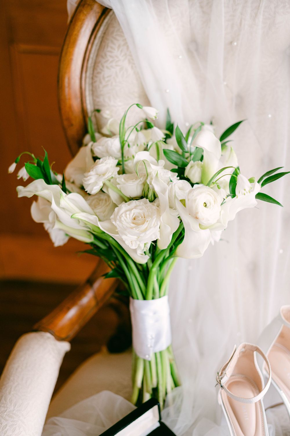 An elegant white wedding bouquet with roses and greenery sits beside blush colored shoes.