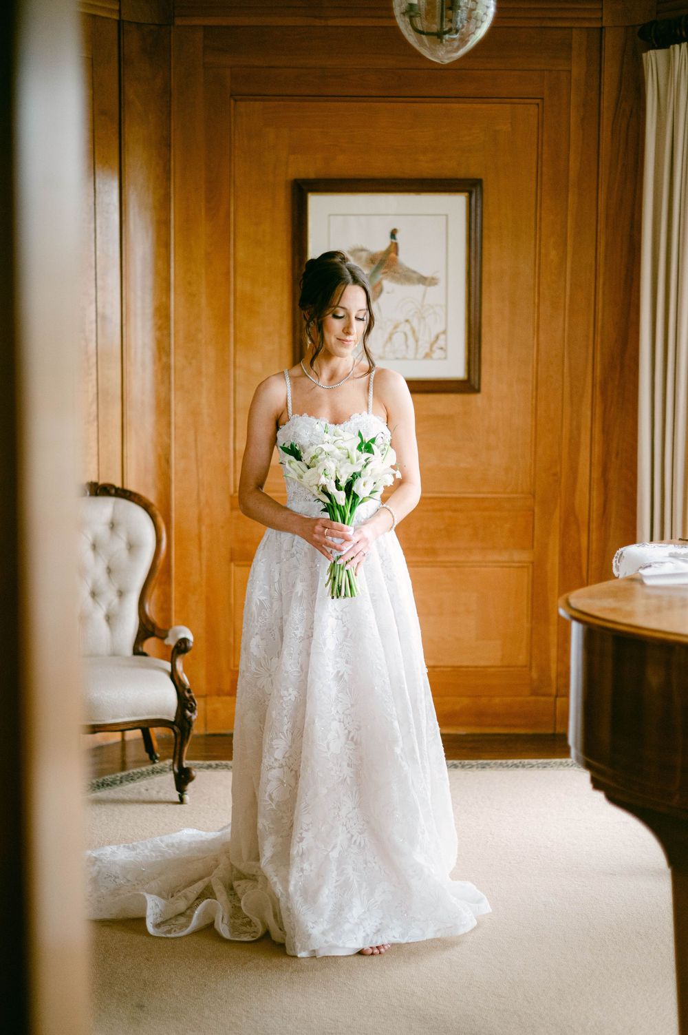 A white lace wedding gown hangs elegantly in front of a sunny window.