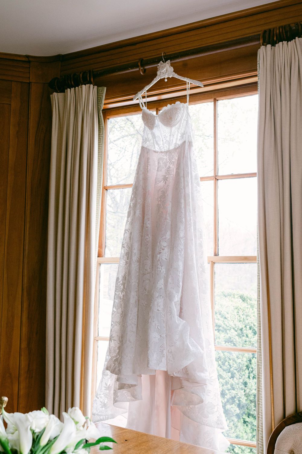 A delicate white lace wedding dress hangs in window light against wooden paneled walls.