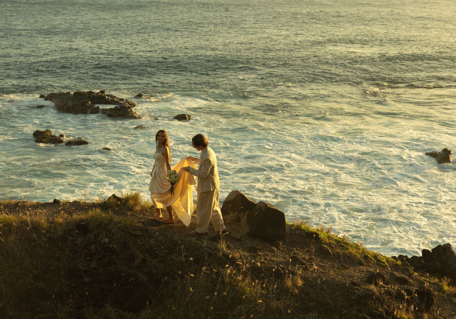 Two figures in flowing dresses stand atop a dramatic coastal cliff at sunset overlooking the ocean waves below.