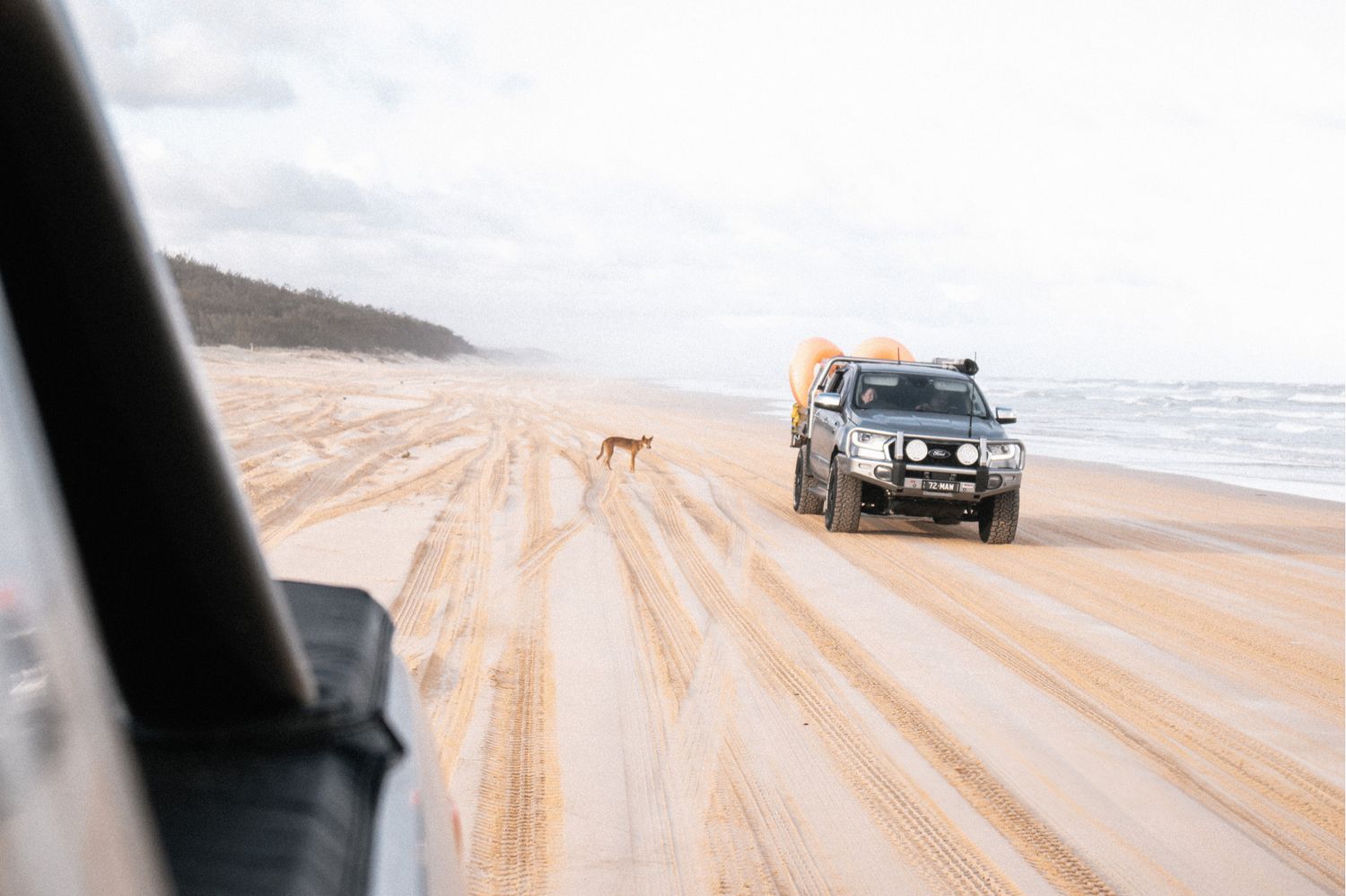 Two off-road vehicles drive along a scenic beach with sandy tire tracks and ocean waves visible on the shoreline.