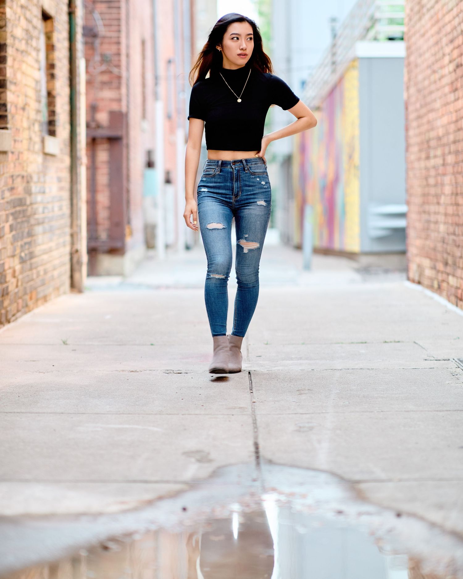 Stylish young woman in a black crop top and ripped jeans posing confidently in an urban alley. Modern lifestyle portrait photography.
