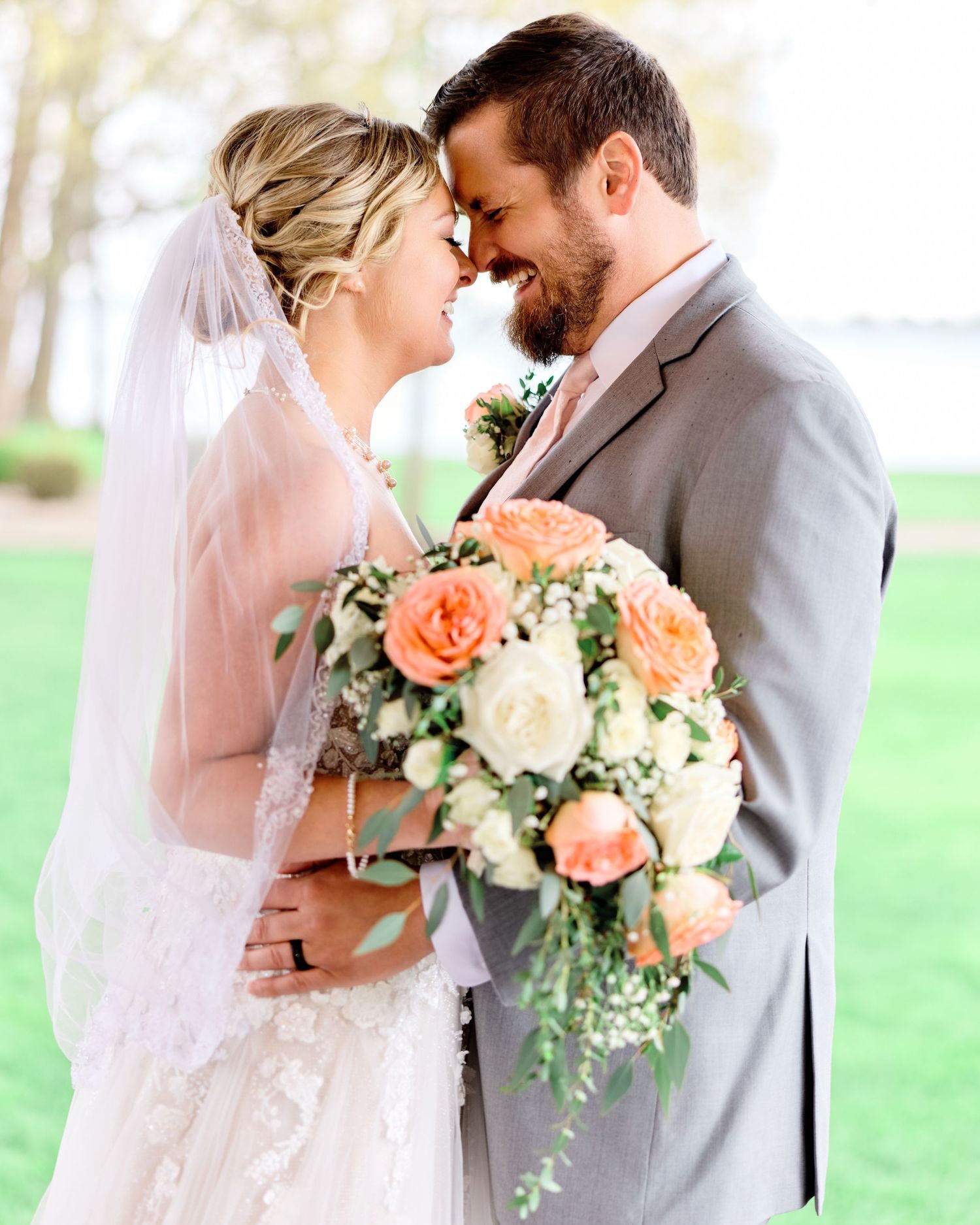 Joyful bride and groom touching foreheads, smiling. Bride holds a peach and white rose bouquet. Romantic wedding photography.