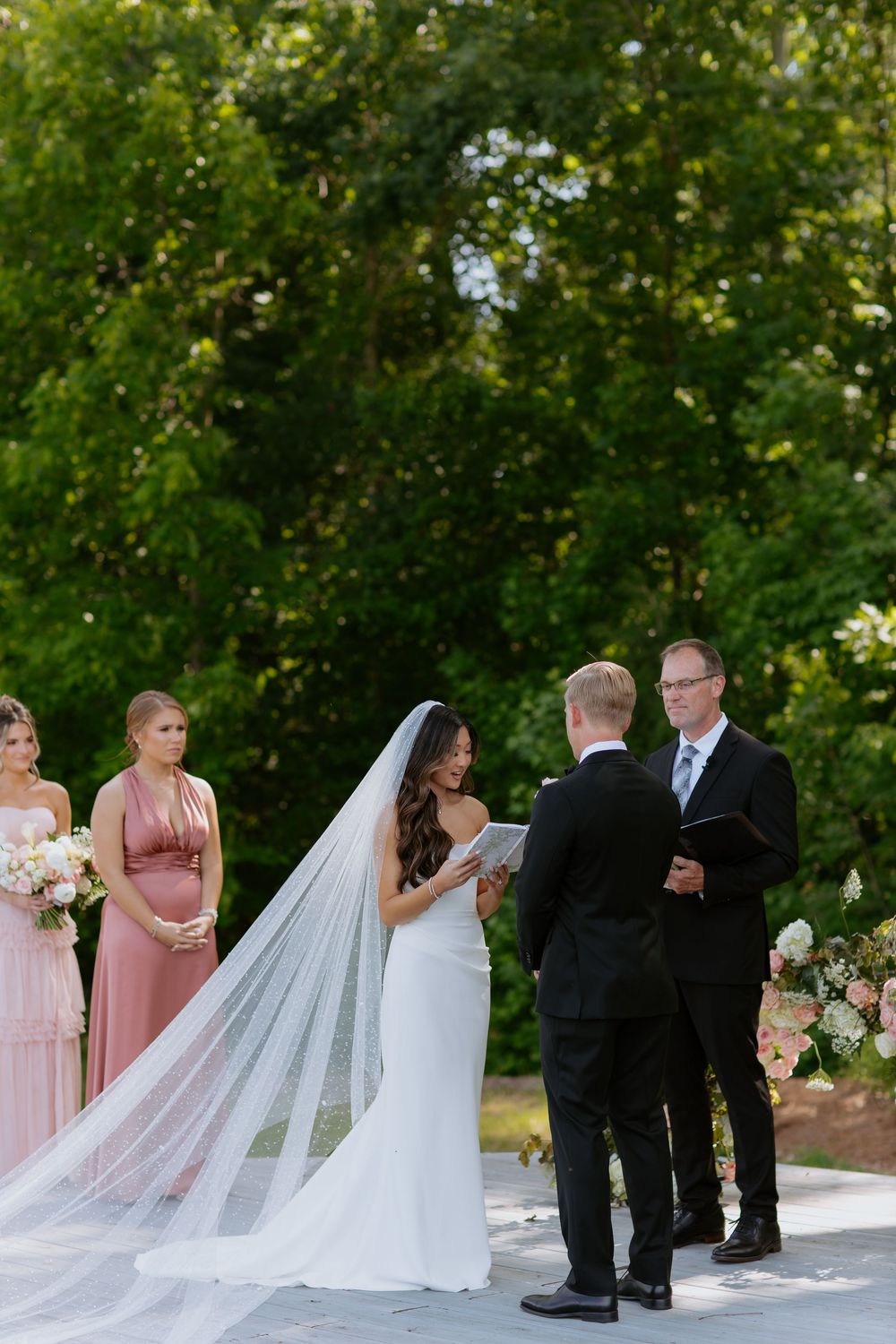 Wedding party lines up outdoors with a flowing white veil in the foreground.
