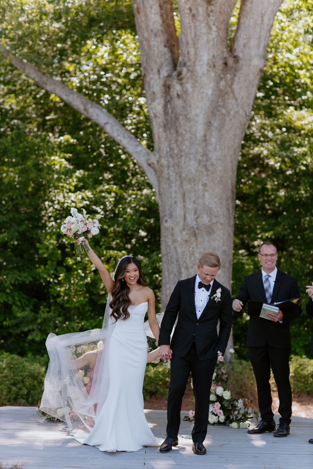 Celebratory wedding ceremony moment on an outdoor platform with large tree in background.