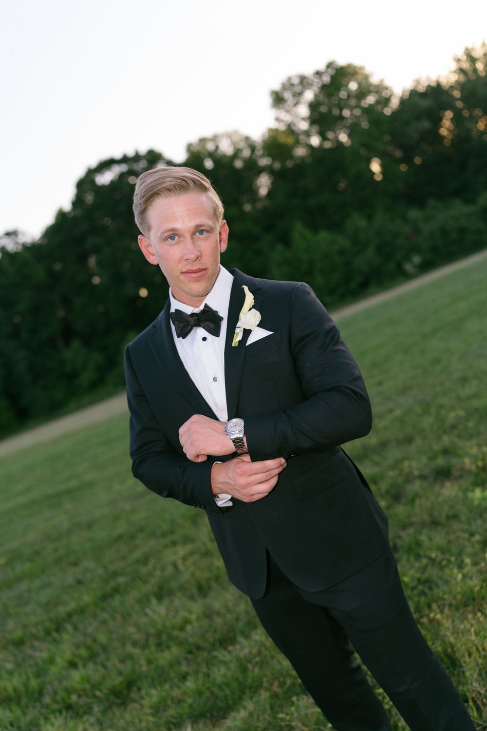 Groom in classic black tuxedo and bowtie adjusts cufflinks on grassy hillside.