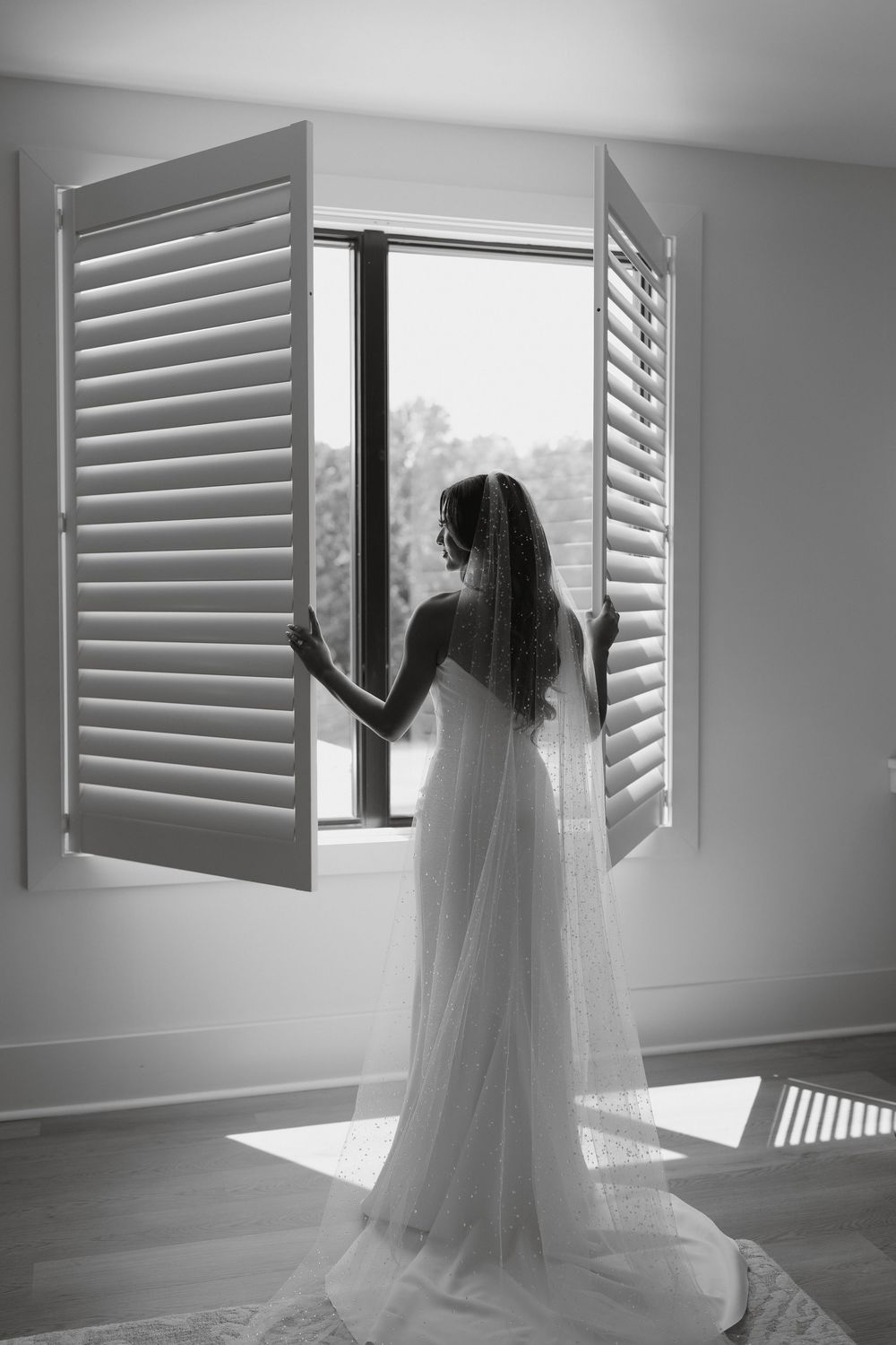 Silhouette of a bride in a flowing wedding gown stands by a window with plantation shutters.