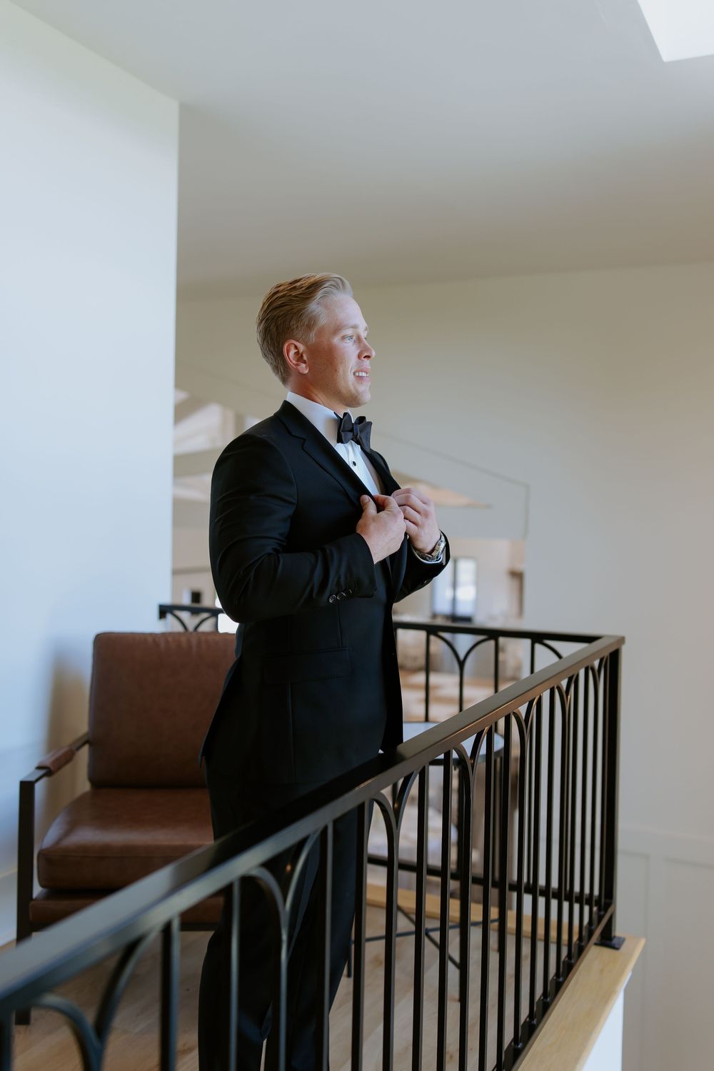 A person in formal attire smiles while standing near a railing in an elegant indoor setting.