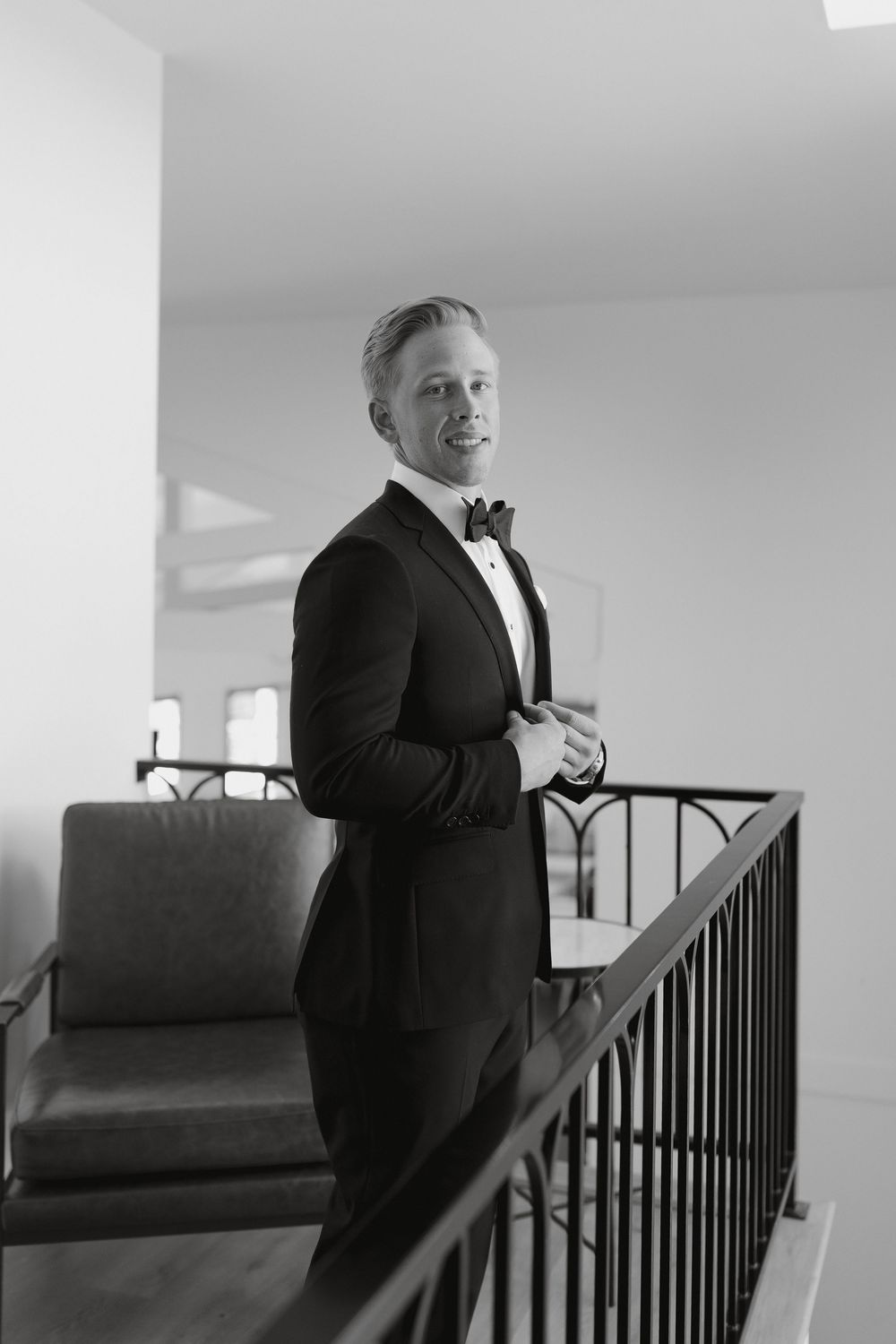 A person in a black tuxedo smiling and adjusting their bowtie while standing on a balcony.