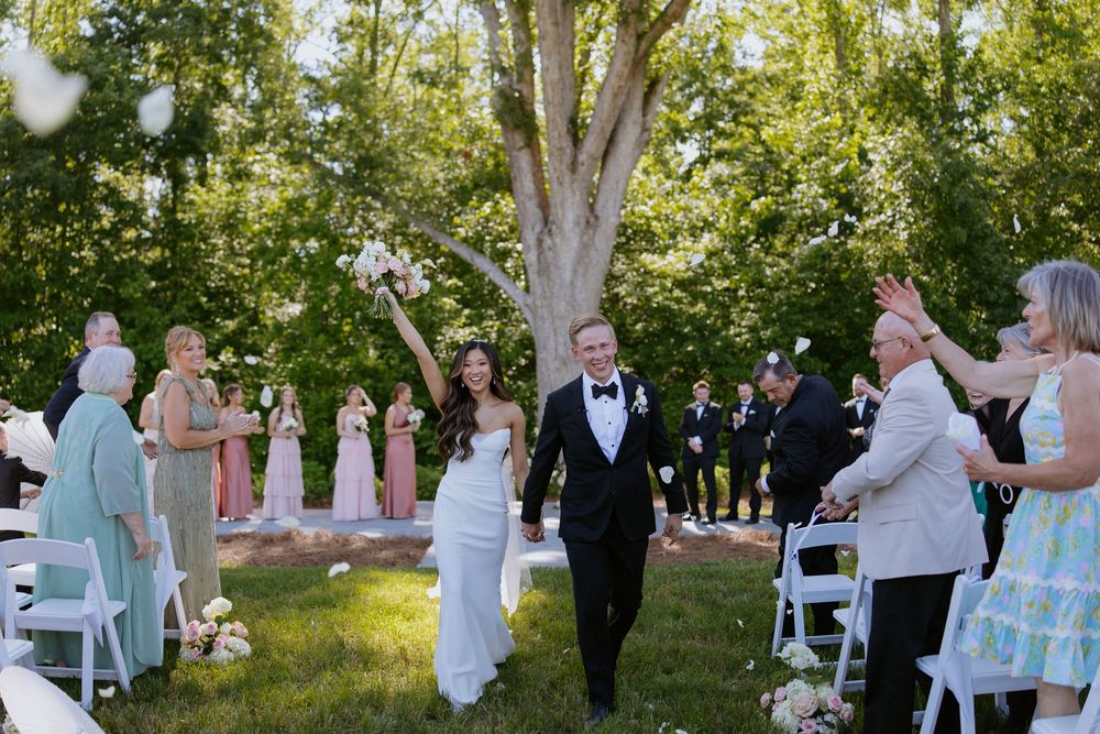 Wedding guests and party members gather outdoors under trees for a ceremony.