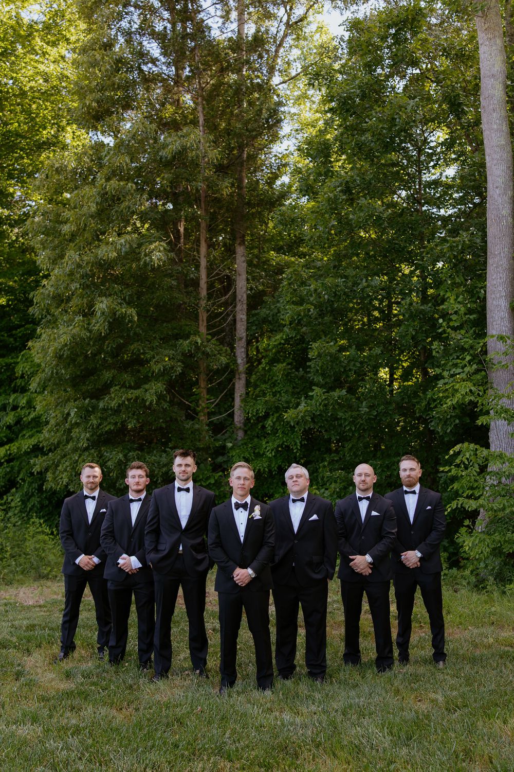 A wedding party in formal black tuxedos stands in line against a backdrop of tall green trees on a grassy lawn.