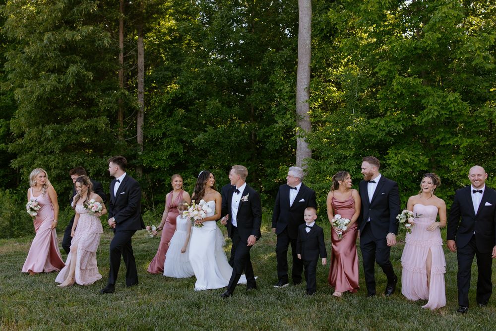 Wedding party in formal attire poses together on grassy lawn with trees in background during golden hour.