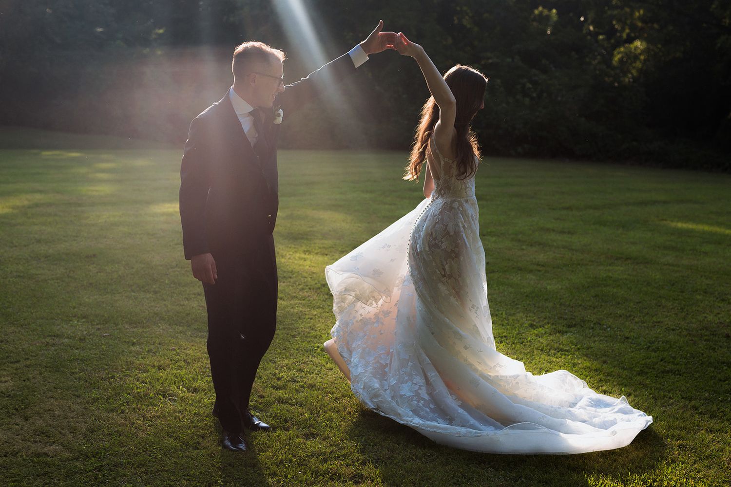 Backlit wedding couple walking across sunlit lawn at golden hour.