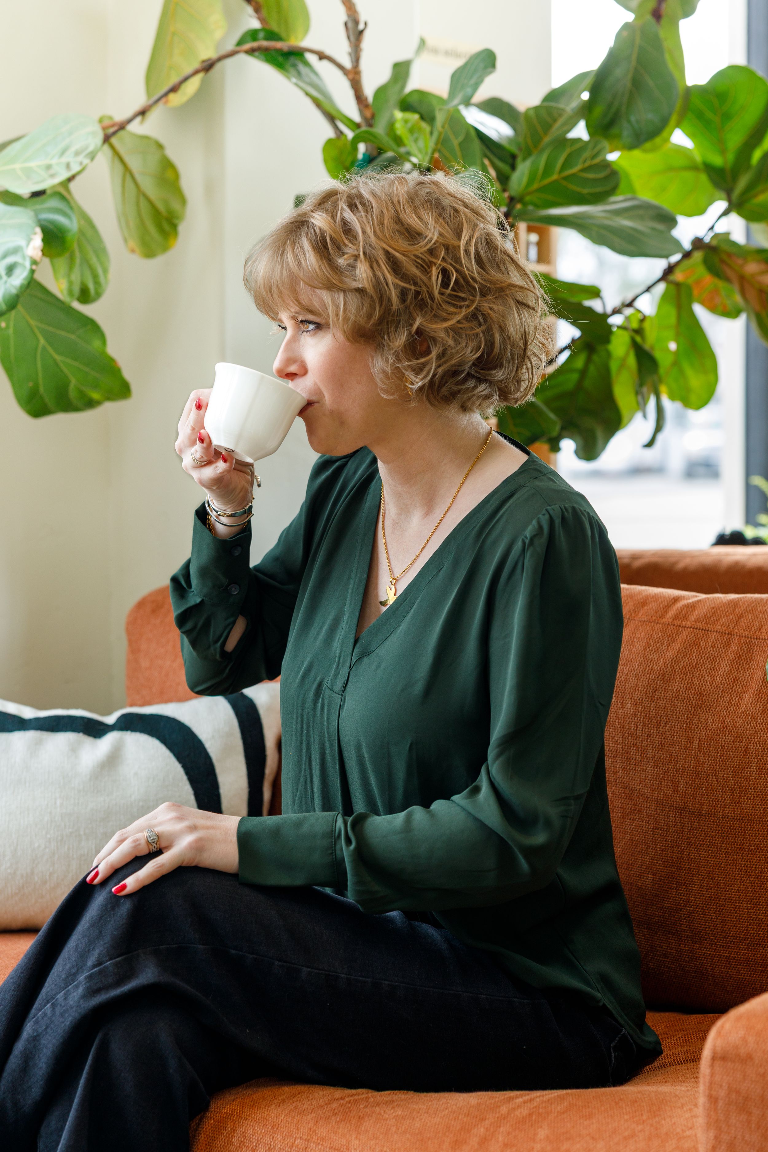 A person in a dark green blouse sips from a white coffee cup while relaxing on an orange couch near indoor plants.