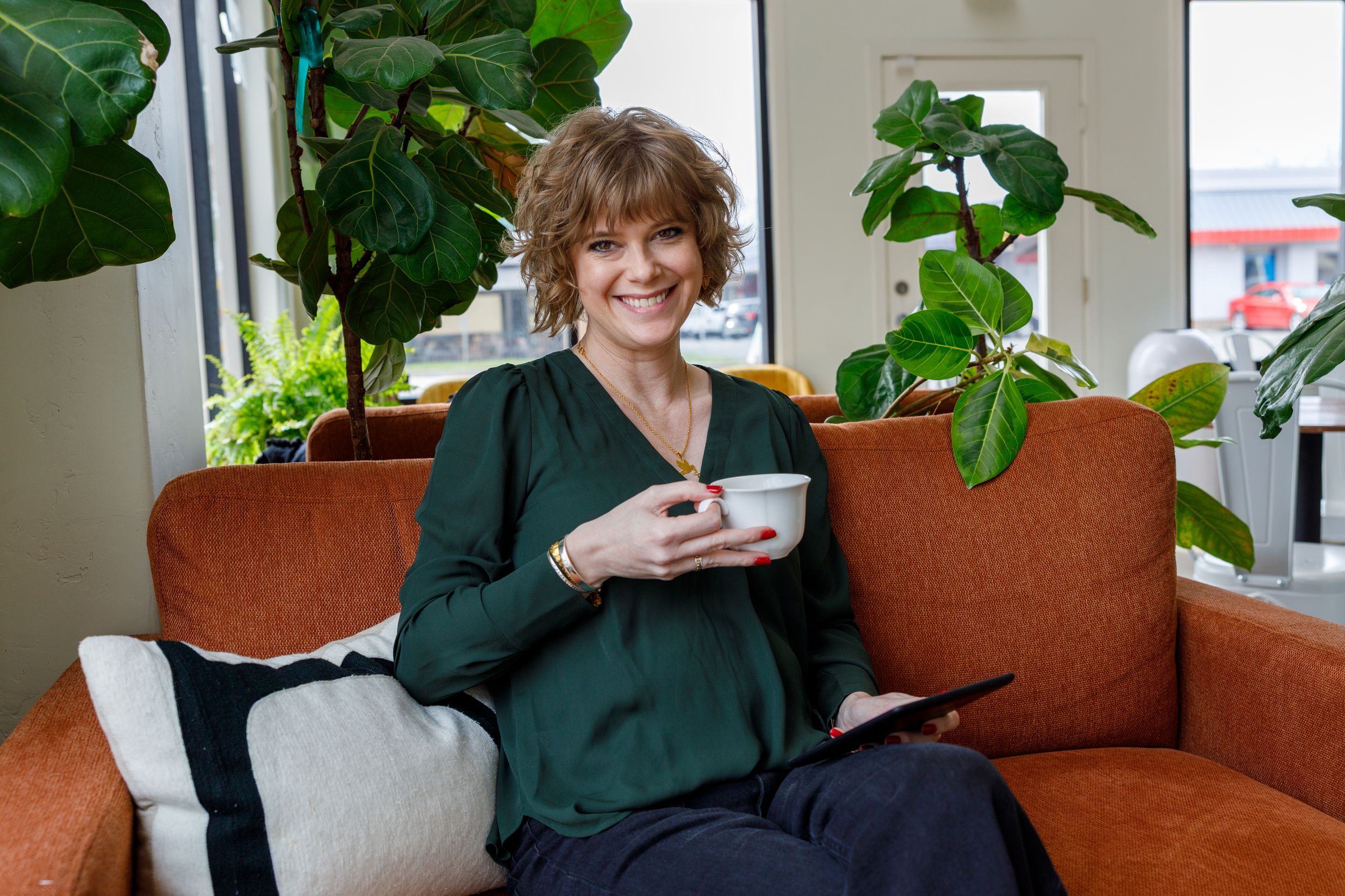 Person relaxes on brown couch with houseplants while holding white mug in green sweater.