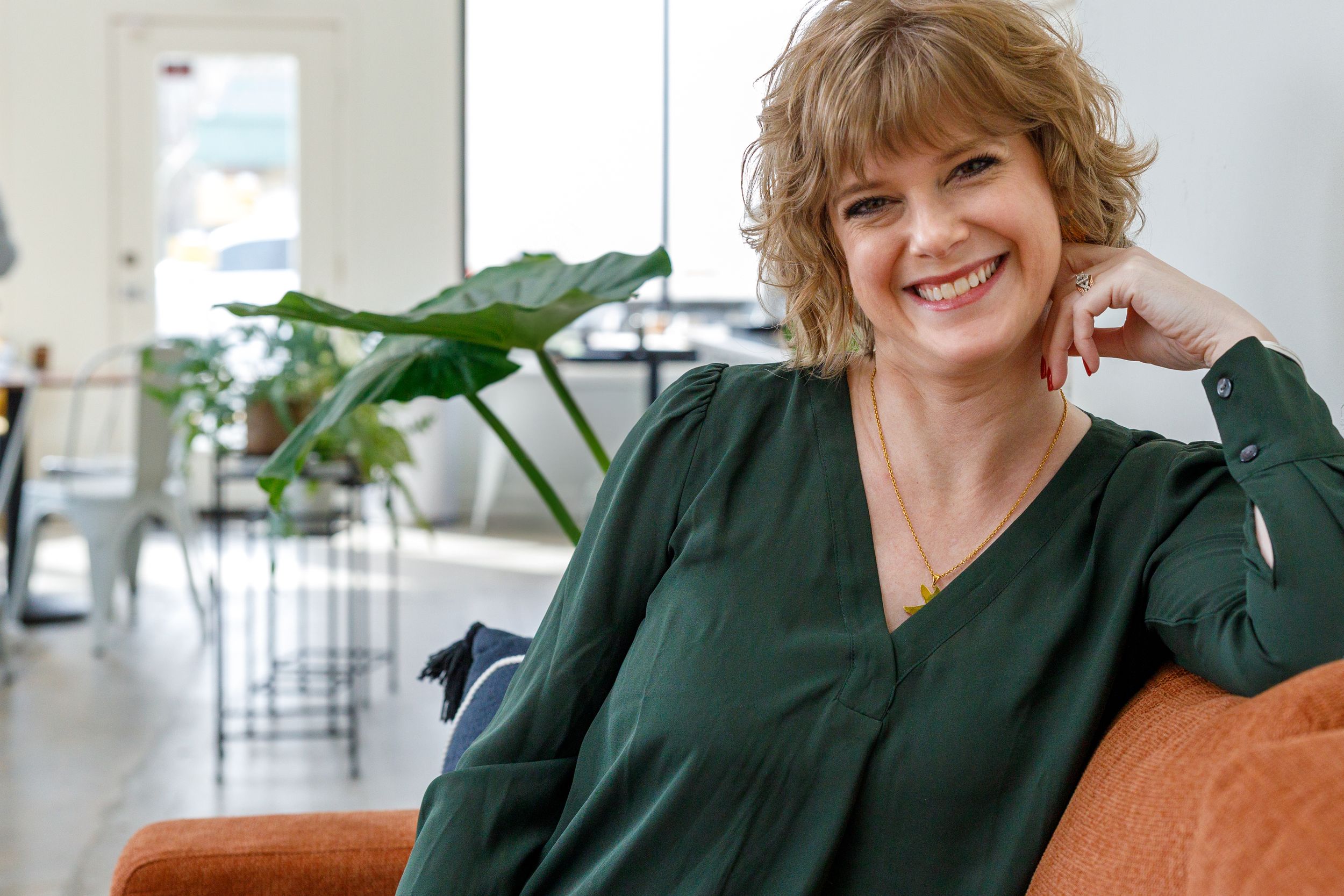 A person in a dark green blouse sits on an orange couch in a bright modern space with indoor plants.
