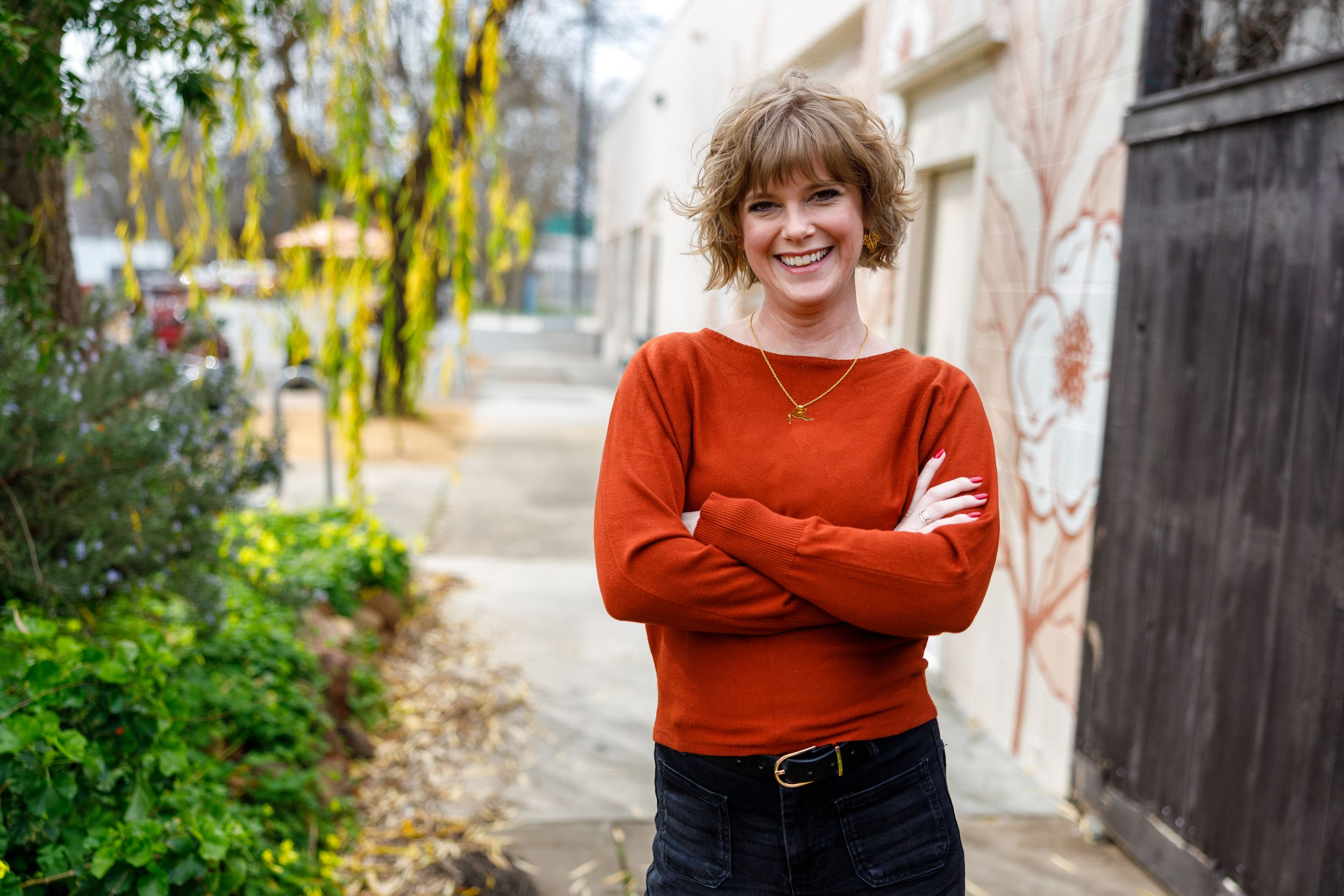 Person in bright orange sweater stands smiling with crossed arms near garden wall outdoors.