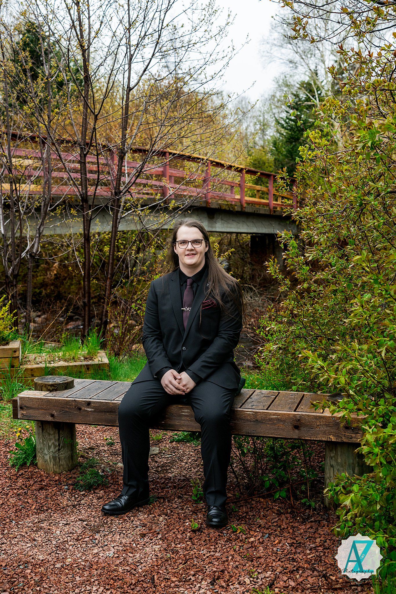 Someone wearing a black suit sits on a wooden bench along a nature trail with a rustic bridge in the background.
