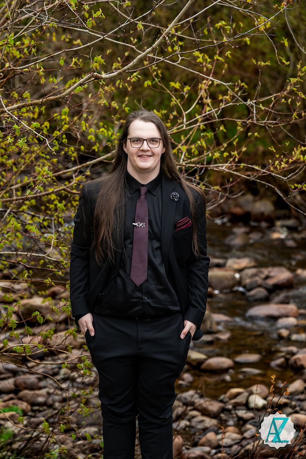 A person in a black suit and purple tie poses outdoors against a backdrop of spring branches and rocky stream bed.