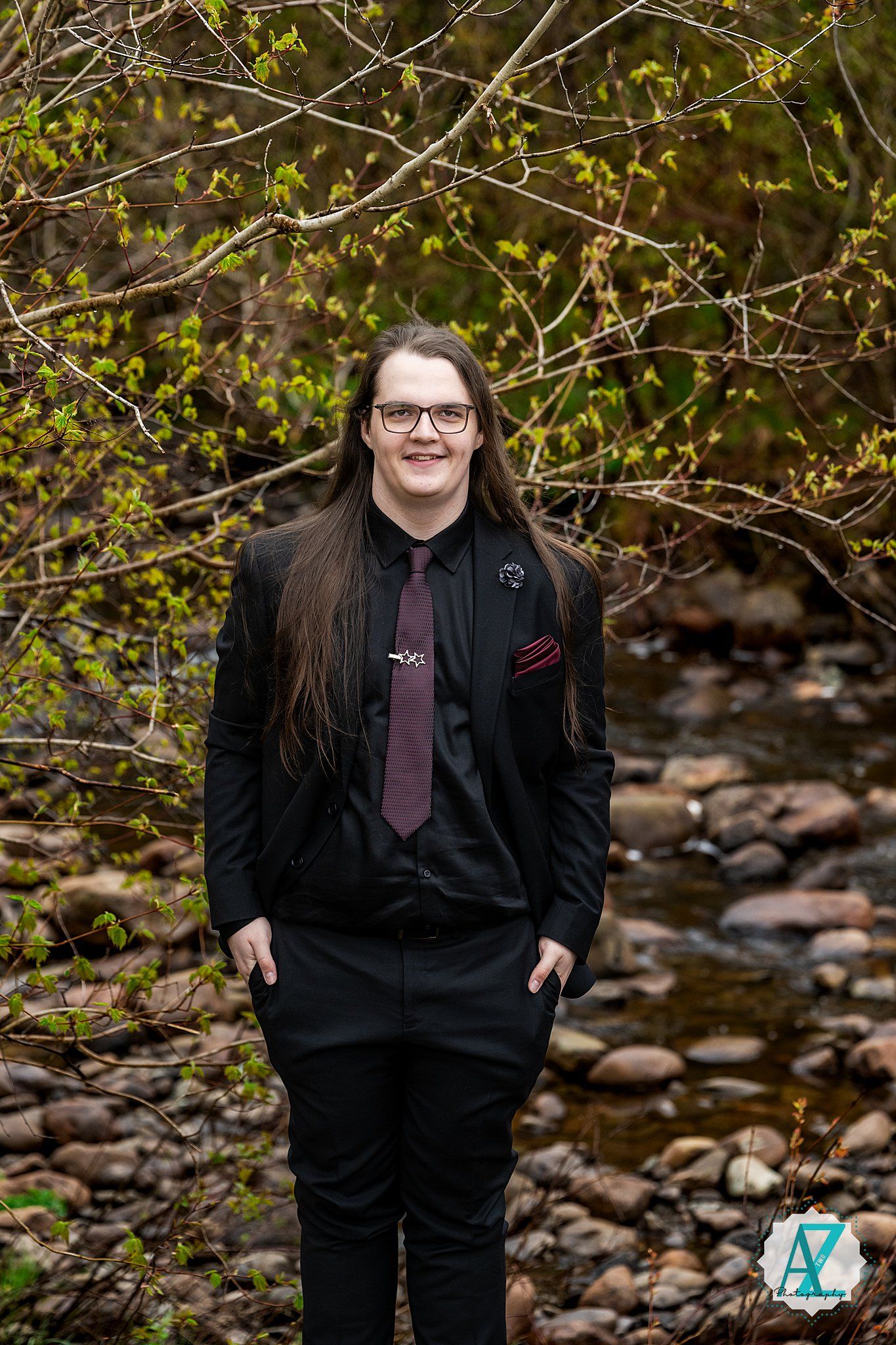 A person in a black suit and purple tie poses outdoors against a backdrop of spring branches and rocky stream bed.