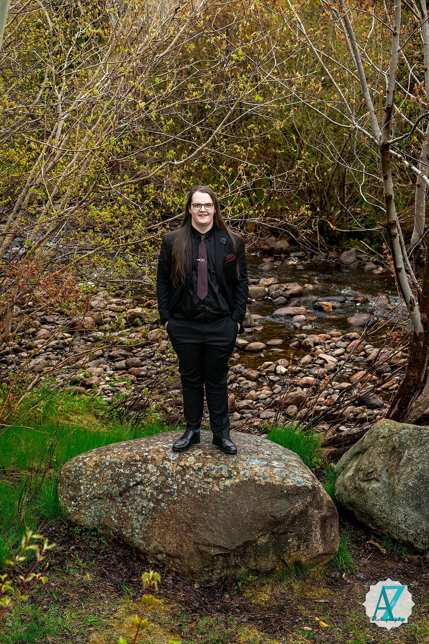 Someone stands on a large boulder near a stream surrounded by autumn foliage and bare branches in a natural setting.