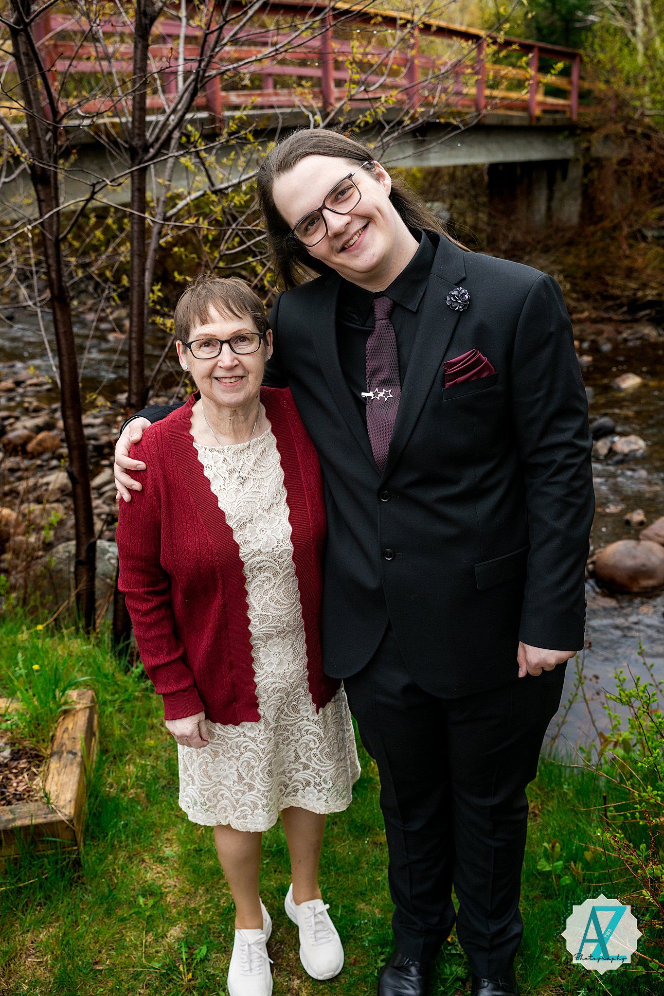 Two people in dressy attire pose in front of a wooden bridge near a creek on a green lawn during springtime.