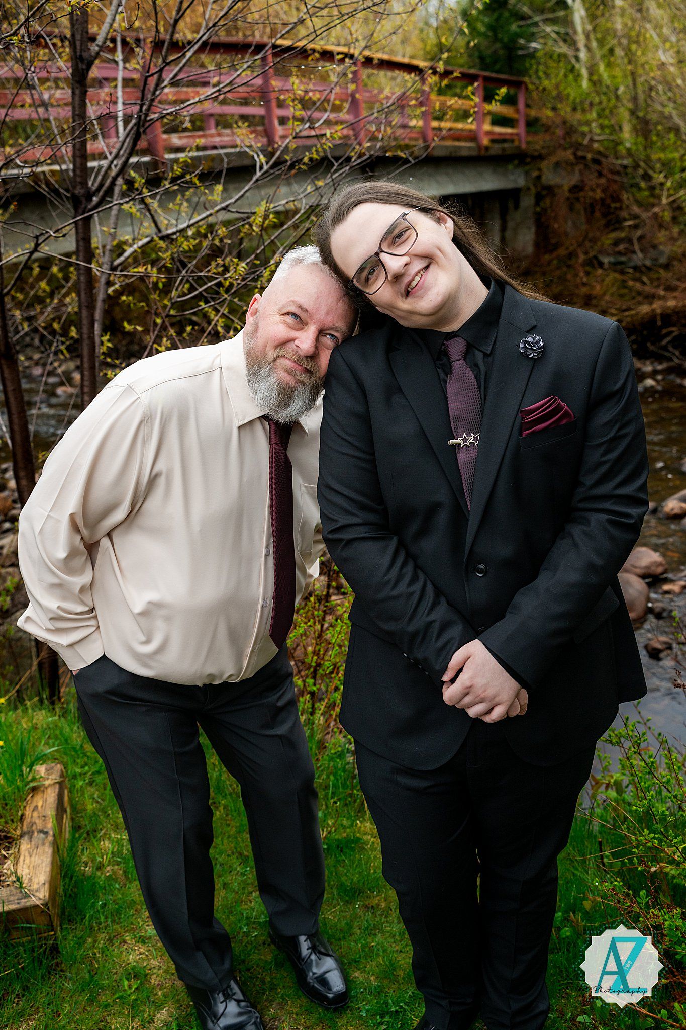 Two people in formal attire pose together on grass with a rustic wooden bridge in the background at twilight.